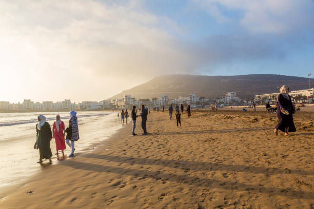 Atmospheric light by the Agadir resort Agadir, Morocco, October 22, 2017: People walk the beach admiring atmospheric sunset light by Atlantic ocean sunset agadir marocco stock pictures, royalty-free photos & images