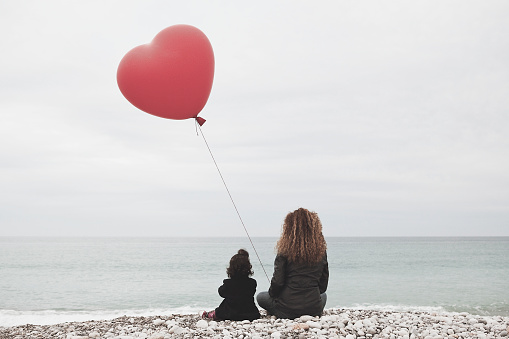 Woman with baby girl and heart balloon looking at the sea