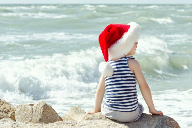 istock Boy in santa hat sitting on a rock. Sea shore. Back view 886625402