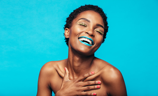 Close up of young woman with colorful makeup smiling against blue background. Beauty shot of african female model with vibrant makeup.