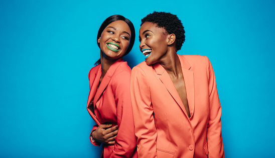 Laughing young women in stylish clothing standing back to back. Female friends standing together in studio over blue background.