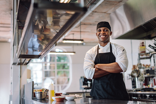 Chef with his arms crossed standing at restaurant kitchen. Male cook wearing apron standing by kitchen counter and smiling.