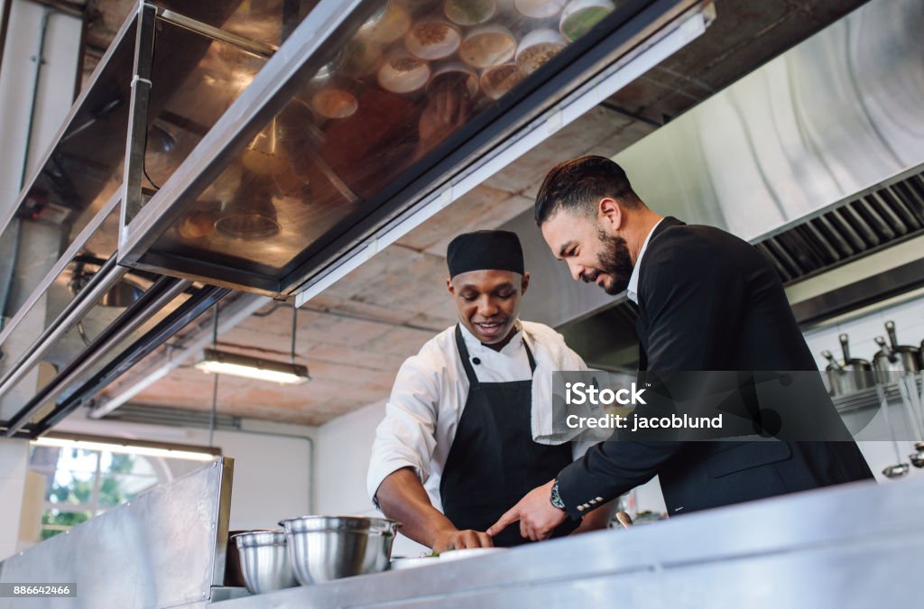 Restaurant manager with chef in kitchen Restaurant manager discussing with chef in kitchen. Cook preparing a dish with restaurant owner standing by. Restaurant Stock Photo