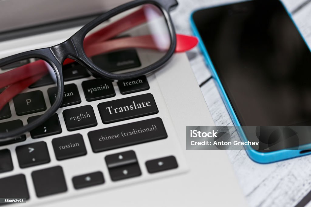 Translation service and application Laptop keyboard with nifty eyeglasses on it and conceptual taglines on the keys - translation to foreign languages concept. Cell phone lies nearby on table. Close-up capture, selective focus. English Language Stock Photo