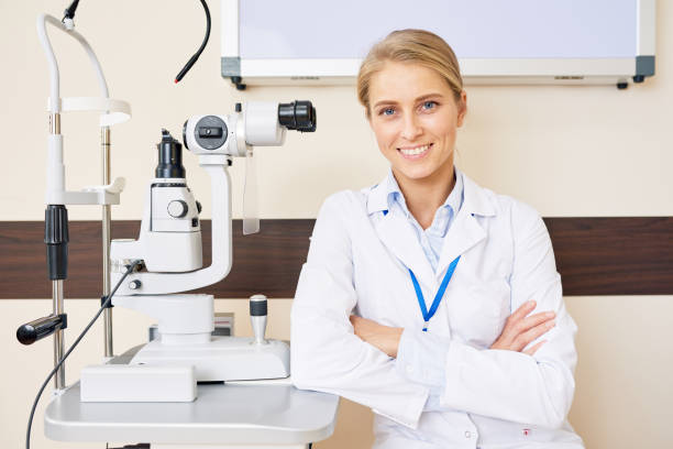 Cheerful Female Ophthalmologist Portrait of blonde female ophthalmologist sitting at slit lamp machine while posing confidently with arms crossed and smiling happily eye test equipment stock pictures, royalty-free photos & images