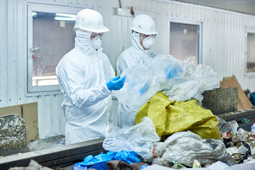 Portrait of two workers wearing biohazard suits at waste processing plant sorting recyclable plastic and cardboard on conveyor belt