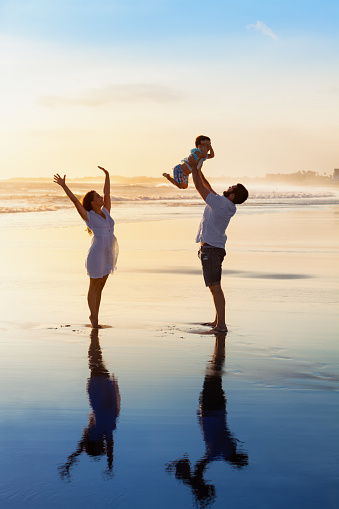 Father tossing high in air baby son, mother jumping by water pool. Happy family walk with fun by sunset black sand beach with sea surf. Active parents, outdoor activity on summer vacation with kids.