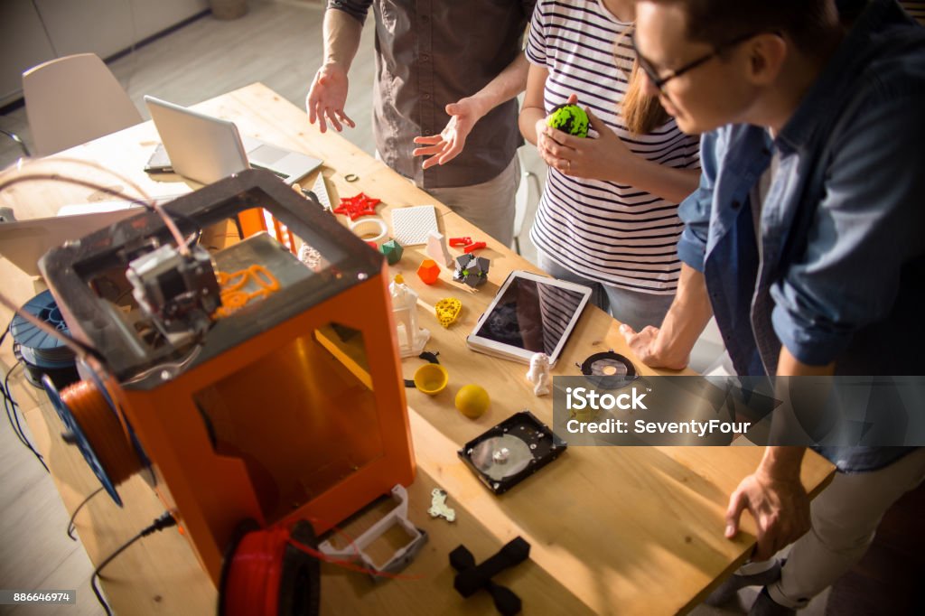 Creative Designers Using 3D Printer High angle of three creative designers working with 3D printer and discussing project plans standing round table with sample plastic models Making Stock Photo