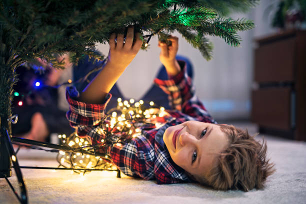Little boy putting lights on a christmas tree. The boy is lying on the floor and attaching the christmas lights to the tree.\n
