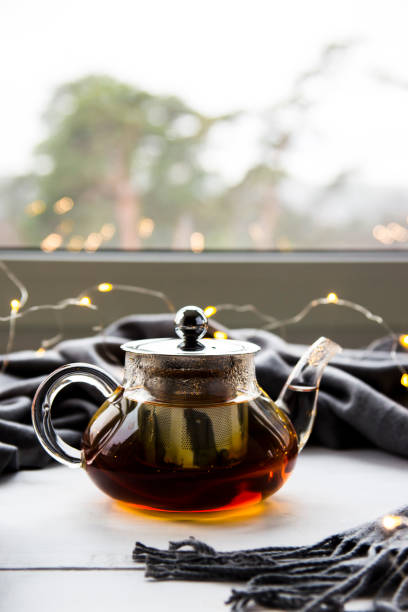 una tetera con té negro al lado de una ventana con una bufanda gris y una guirnalda de la navidad. concepto de invierno - texas tea fotografías e imágenes de stock