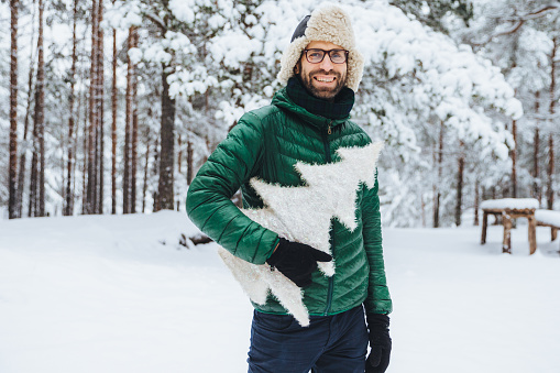 Pleasant looking bearded male wears warm clothes, holds white fir tree, stands against winter forest background, walks with friends during frosty weather, being in good mood. People and nature