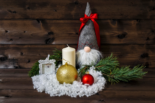 Christmas decoration with pine branches, white candle, picture frame, shiny Christmas balls, snow & scandinavian traditional gnome tomte, nisse, tomtenisse, tonttu with red ribbon on wooden background.