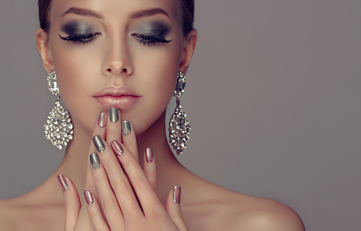 Beautiful woman-model dressed in a big silver earings is showing silver and rose manicure on the nails and rose lipstick on the lips. Fashion makeup and cosmetic.