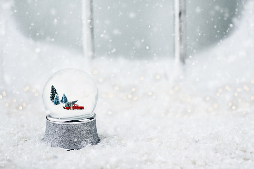 Silver snow globe with antique toy truck hauling a Christmas tree. Snowglobe is sitting outdoors on the ledge of an old wooden window in the snow.