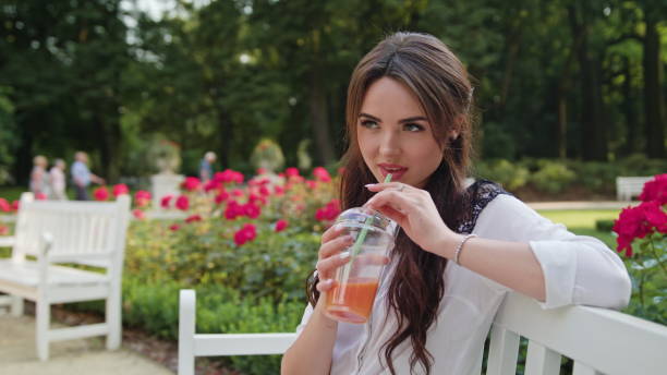 istock Brunnette Lady Sitting on a Bench in the Park Drinking 886933850