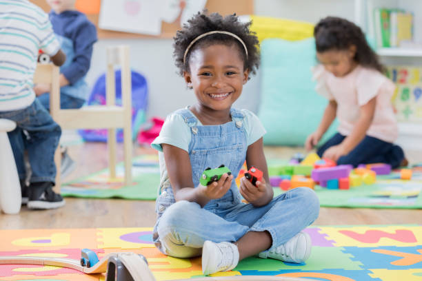 Adorable little girl enjoys time at day care An adorable preschool age girl sits on the floor of her day care classroom and smiles for the camera.  She is sitting beside a toy train track and holding two wooden train cars. miniature train stock pictures, royalty-free photos & images