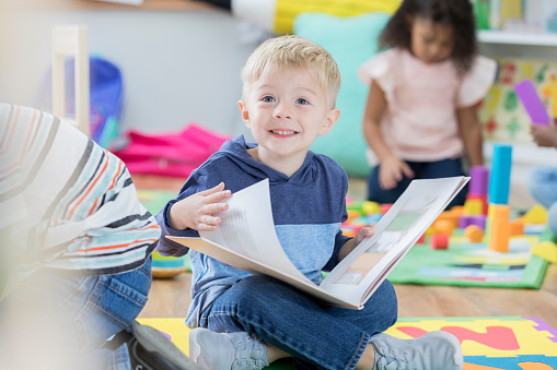 An attentive cheerful preschool age boy sits on the floor of his classroom during free play time and looks up.  He is distracted from turning a page in a picture book.