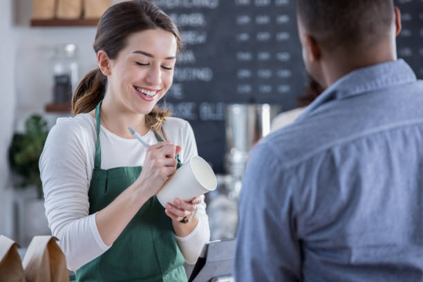 cheerful coffee shop barista writes order on cup - coffee serving cafeteria worker checkout counter imagens e fotografias de stock