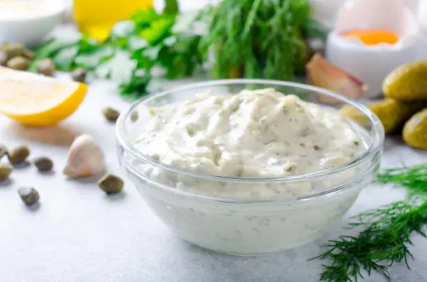 Homemade white sauce tartar tartare with ingredients pickles, capers, dill, parsley, garlic, lemon and mustard on a light stone background. Horizontal image