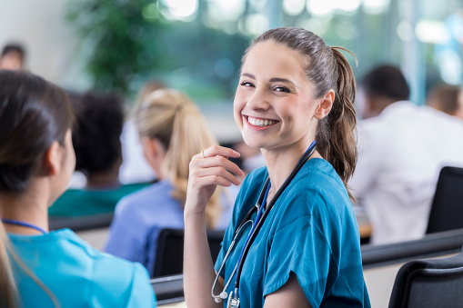 Cheerful young Hispanic nursing student smiles confidently before class. She is sitting next to her friend. Other classmates are in the background.