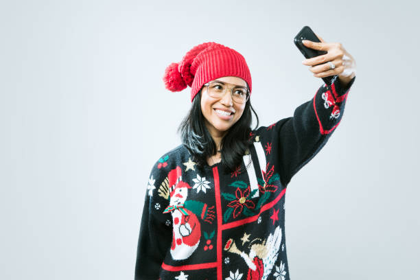 Christmas Sweater Woman Taking Selfie A happy Asian - Pacific Island woman wears an ugly Christmas sweater, having fun during the holiday season.  She takes a self portrait with her smart phone to share on social media. christmas sweater stock pictures, royalty-free photos & images