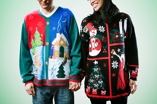 A man and woman wear ugly Christmas sweaters, the woman having fun and the man not enjoying it that much.  Horizontal image with green background.