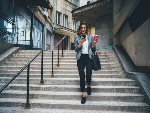 Woman texting and drinking coffee outdoors Woman texting and drinking coffee as she's walking down the stairs in the city. smart casual stock pictures, royalty-free photos & images