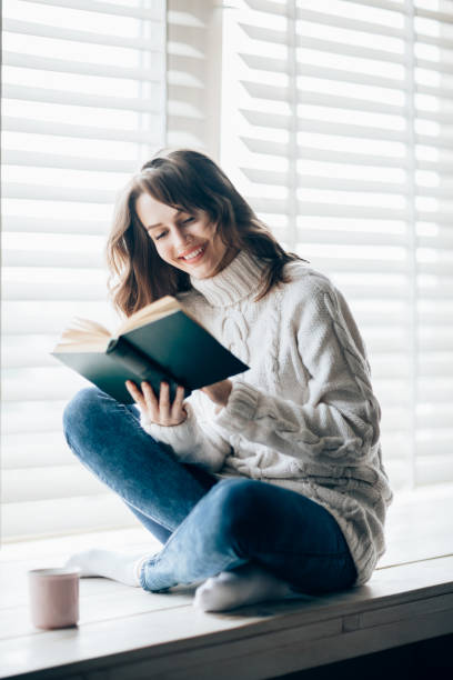 istock Happy beautiful woman reading book sitting on window sill 886949128