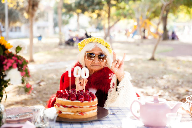 older woman with a gift wear yellow sweater and horn cap on a white background holding plate with cake with fireworks.