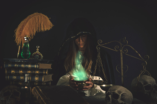 Woman wizard at an old alchemists work table with mystical items, books, potions, skulls, scales and other equipment.