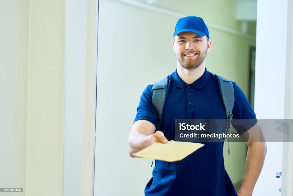 Home mail delivery Positive friendly young postman in cap giving letter to customer Envelope Stock Photo
