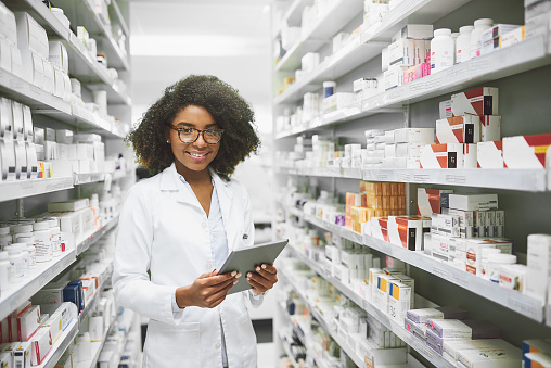 Portrait of a cheerful young female pharmacist standing with a digital tablet while looking at the camera in a pharmacy