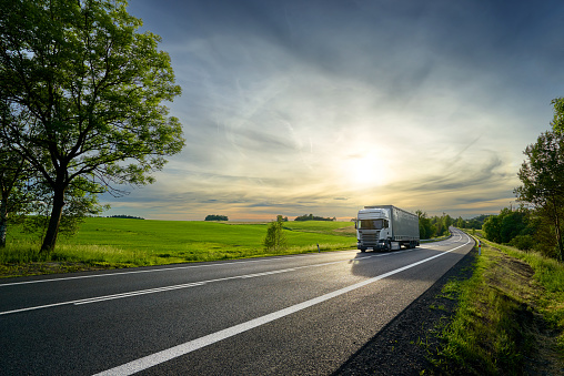White truck driving on the asphalt road next to the green field in rural landscape at sunset