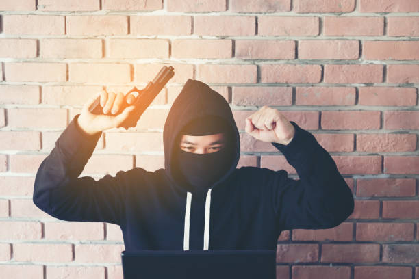 Hacker holding gun working on his computer, war, terrorism , terrorist and bandit concept. Glitch effect and copy space. Black masked hacker in the black long sleeve shirt is sitting on the desk in front of laptop. hacker con pistola trabajando en su concepto de equipo, guerra, terrorismo, terrorista y bandido. falla efecto copia espacio. negro enmascarado hacker negro camisa de manga larga está sentado en el escritorio delante del ordenador portátil.