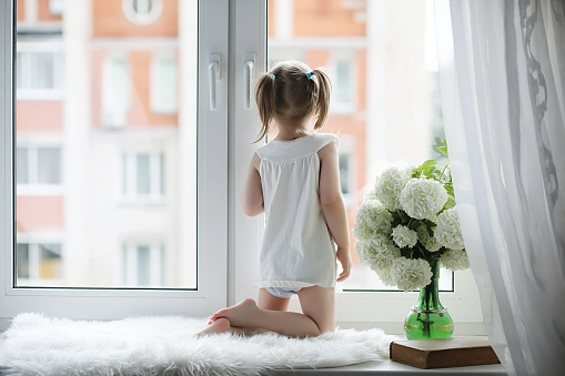 A little girl is sitting on the windowsill. A bouquet of flowers in a vase by the window and a girl sniffing flowers. A little princess in a white dress with a bouquet of white