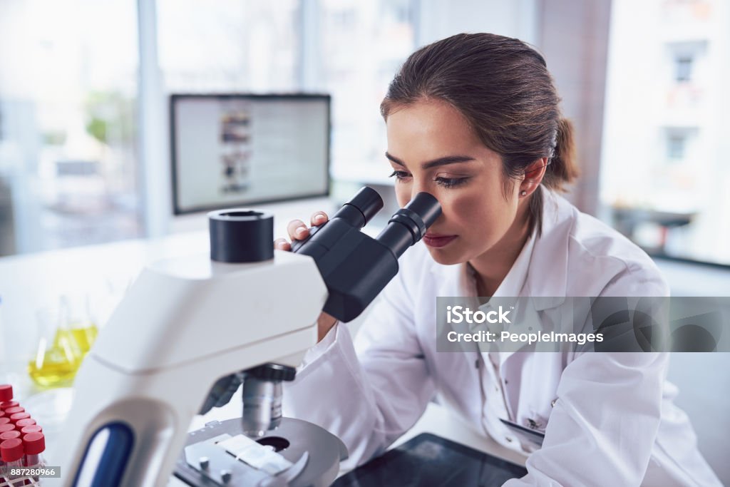 State of the art science equipment Shot of a cheerful young female scientist looking through the lens of a microscope while being seated inside of a laboratory Laboratory Stock Photo