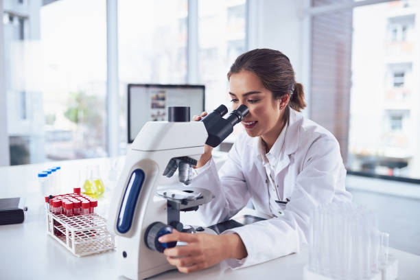 She stumbles across new findings everyday Shot of a cheerful young female scientist looking through the lens of a microscope while being seated inside of a laboratory biochemist photos stock pictures, royalty-free photos & images