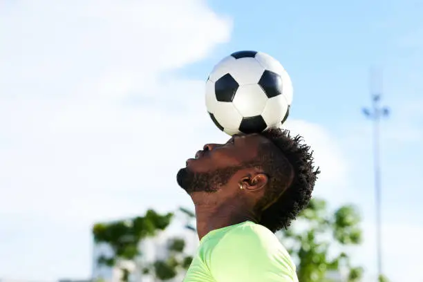Profile view of concentrated African soccer player balancing ball on head while having training outdoors, blue sky on background