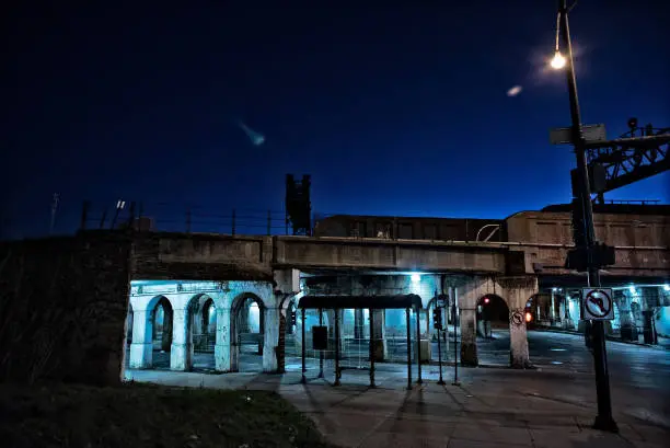 Photo of Gritty dark Chicago city street intersection under industrial train bridge viaduct tunnel with a CTA bus station at night.