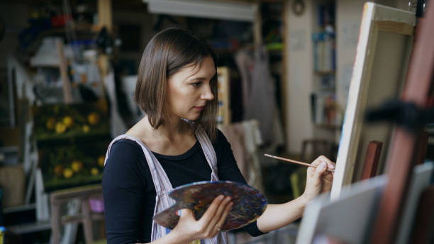 Young woman artist in apron painting picture on canvas in art studio Young woman artist in apron painting picture on canvas in art studio indoors abstract oil painting for beginners stock pictures, royalty-free photos & images