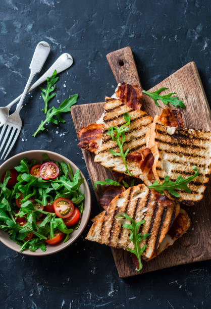 pancetta alla griglia, panini alla mozzarella su taglieri e rucola di legno, insalata di pomodoro ciliegino su sfondo scuro, vista dall'alto. deliziosa colazione o spuntino, laici piatti - mozzarella tomato sandwich picnic foto e immagini stock