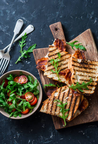 グリル ベーコン、モッツァレラチーズの木製のまな板とルッコラ、暗い背景、上面にチェリー トマトのサラダのサンドイッチします。おいしい朝食やおやつ、フラット レイアウト - mozzarella tomato sandwich picnic ストックフォトと画像
