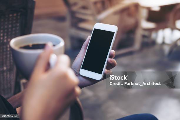 Mockup Image Of Womans Hands Holding White Mobile Phone With Blank Black Screen While Drinking Coffee In Modern Loft Cafe Stock Photo - Download Image Now