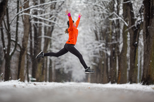active woman in running outfit jumping during snowfall in winter december in alley snow on road and trees cold bad weather