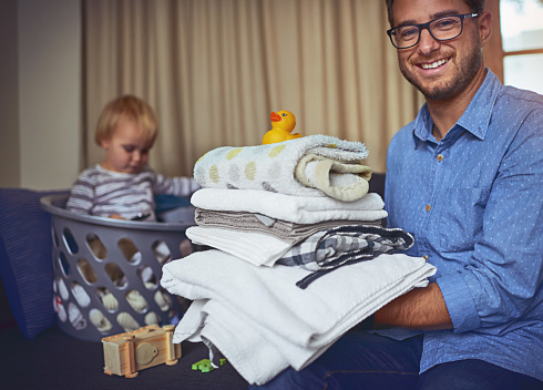 Portrait of a young man holding a load clean laundry with his son in the background