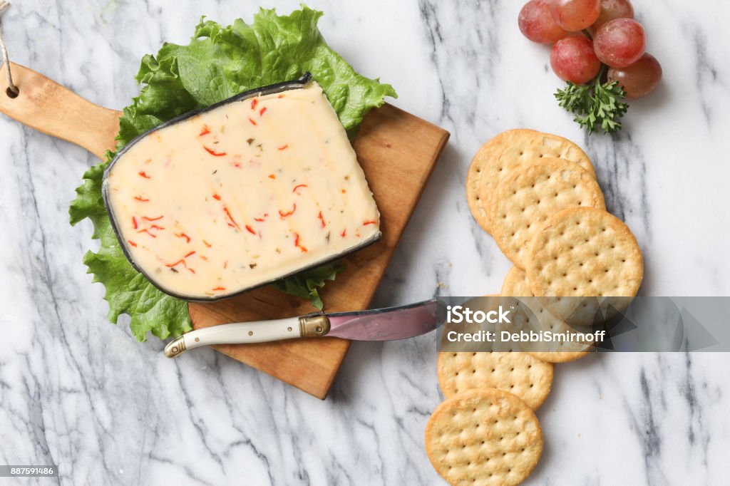 Artisanal Cheese And Crackers An overhead, close up horizontal photograph of a wedge of Jalapeño/Piquanté Peppersflavored artisanal cheese on a small wooden cutting board, some crackers and a knife. Garnished with some green lettuce and red grapes. Artisanal Food and Drink Stock Photo