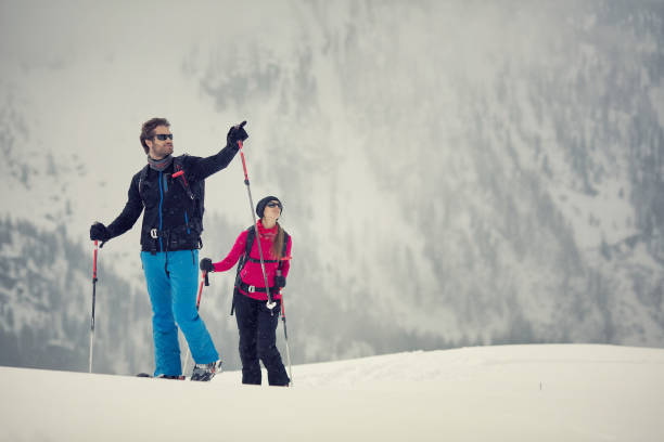 couple man and woman skier exploring snowy land walking and skiing with alpine ski. europe alps. winter sunny day, snow, wide shot, warm sun flare.travelling - summit cross imagens e fotografias de stock