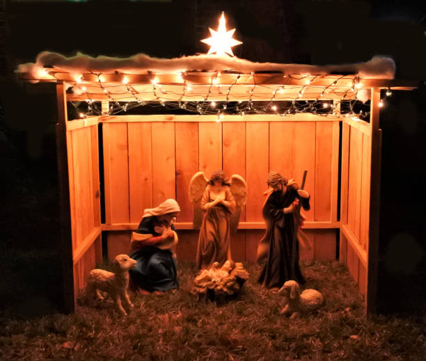Large Outdoor Christmas Nativity Creche A large wooden creche illuminated by string lights and a star is set up on a front yard at night showing an angel visiting Mary and Joseph and two sheep as Jesus is born and lays in a manger in a stable. lawn nativity scene stock pictures, royalty-free photos & images
