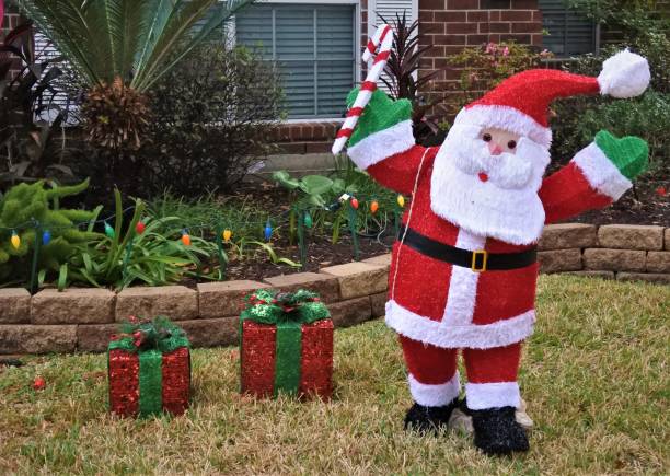 A jolly statue of Santa Claus raises a candy cane beside two gifts on a front lawn.