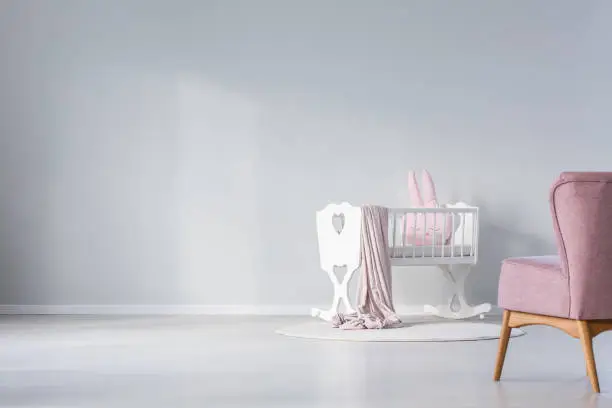 Copy space of empty wall in baby's room with pink rabbit cushion in white cradle near rug and violet chair